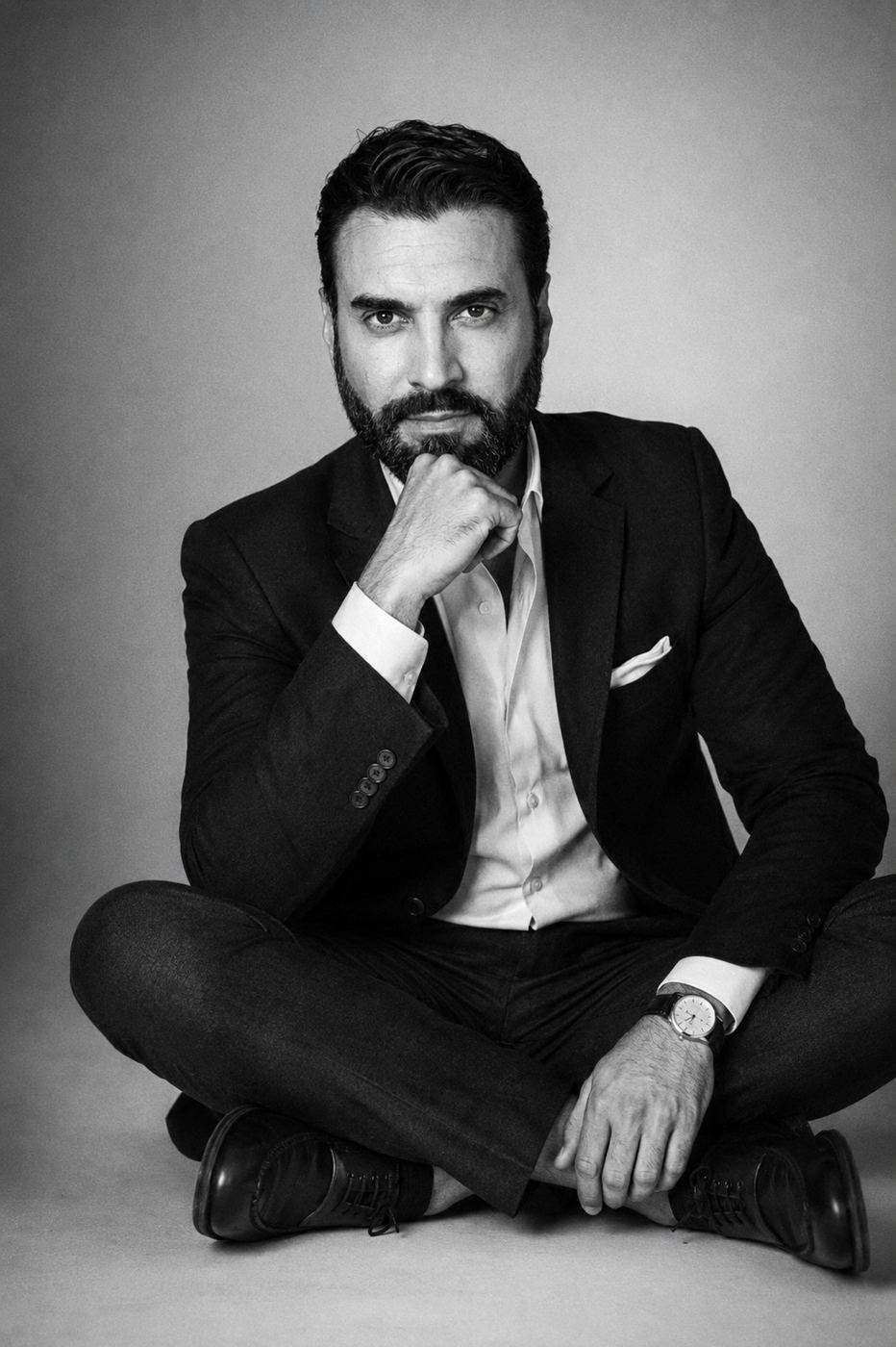 A.J. Castro seated cross-legged in a suit, black and white
